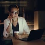 Woman in glasses working late at night on laptop, holding coffee, making phone call.